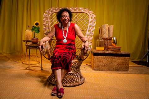 Mulher negra de cabelos curtos e cacheados sorri, sentada em uma grande poltrona de vime trançado. Ela veste roupa vermelha com estampas pretas, usa colares compridos e sandálias vermelhas. O cenário, em tons de amarelo e dourado, é composto por cortinas, tapete e móveis de fibras naturais. Nas laterais, pequenos arranjos decorativos incluem um girassol, vasos, garrafas e cestos.