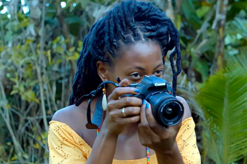 Cena do curta-metragem Como Matar uma Boneca. Em meio à vegetação, uma jovem negra de cabelos longos em tranças segura uma câmera fotográfica Canon diante do rosto, preparando-se para fotografar. Ela veste uma blusa amarela de ombros descobertos e usa brincos compridos.