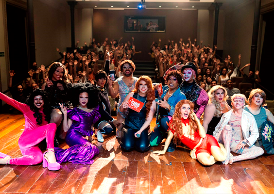 Fotografia de um grupo de drags queens, com vestimentas coloridas e brilhantes, sentadas em um palco de madeira. Atrás dela, uma plateia cheia com as mãos levantadas para cima. Todos parecem animados.