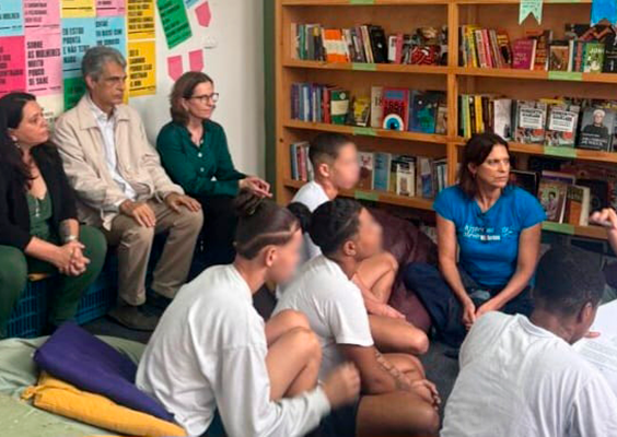 A imagem mostra um grupo de pessoas em uma sala de leitura. Na parte de trás, algumas pessoas adultas estão sentadas em bancos e cadeiras próximas a uma estante cheia de livros. Na frente, crianças ou adolescentes estão sentados no chão, de costas para a câmera, aparentemente ouvindo uma pessoa que segura um papel. A parede ao fundo está decorada com cartazes coloridos e escritos. A cena parece retratar um momento de aprendizado ou leitura em grupo, em um ambiente acolhedor e educativo.