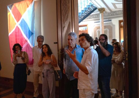 A imagem mostra um grupo de pessoas em um ambiente interno com decoração sofisticada. À esquerda, uma bandeira colorida com formas geométricas está pendurada na parede. No centro, um homem de camisa branca faz um gesto explicativo enquanto outros escutam, incluindo uma mulher segurando um celular e um homem mais velho de camisa azul. Ao fundo, uma porta aberta revela colunas e detalhes arquitetônicos elaborados. O clima é de interação e atenção.
