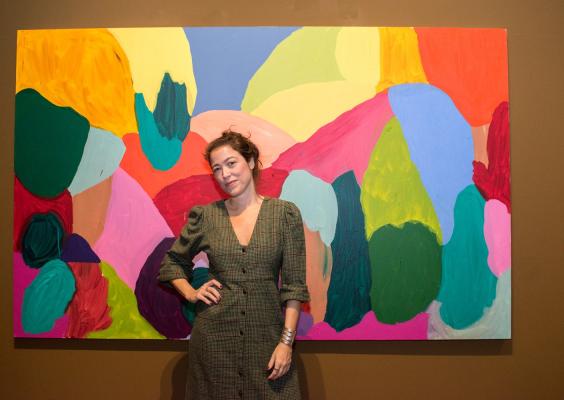 A imagem mostra uma mulher de vestido xadrez na frente de um quadro grande com formas abstratas de cores vivas. Ela é branca, de cabelos escuros e presos. Está sorrindo e com uma das mãos na cintura.