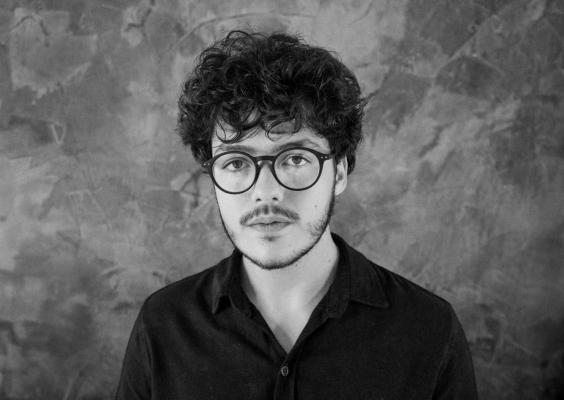A foto em preto e branco mostra um homem jovem, branco. Ele possui cabelos enrolados escuros e barba cerrada. Usa óculos de grau redondo, com armação escura. Veste camisa de gola preta. Olha fixamente para a câmera. 