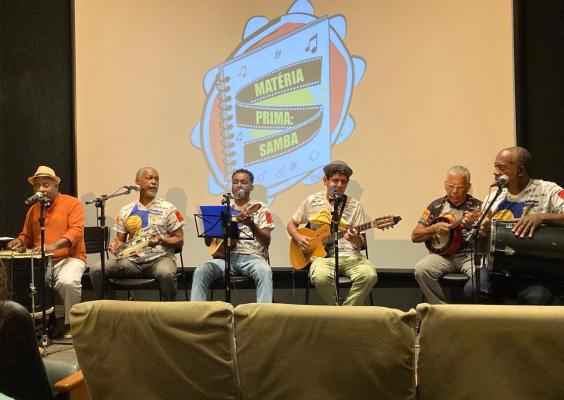 A imagem mostra um grupo de seis músicos tocando em um palco, provavelmente em um evento dedicado ao samba. Eles estão sentados em frente a uma tela de projeção que exibe o título “Matéria Prima: Samba” dentro de uma ilustração de pandeiro. Cada músico está com um instrumento típico do samba, como pandeiro, cavaquinho, violão e tantã, e alguns deles cantam ao microfone. A cena transmite um clima de descontração e celebração da música brasileira, especialmente do samba.
