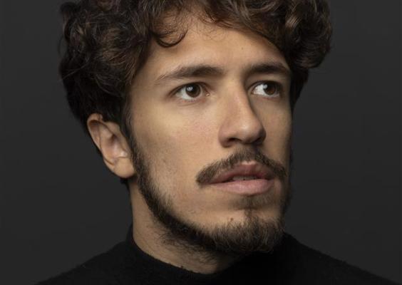 A imagem mostra um retrato de um homem jovem com cabelo ondulado e barba curta. Ele veste uma blusa preta de gola alta e está posicionado contra um fundo escuro, que destaca seu rosto. O olhar do homem está direcionado levemente para a esquerda, e sua expressão é contemplativa, dando um ar sério e artístico à foto. A iluminação é suave e enfatiza as feições do rosto, criando um contraste elegante com o fundo.