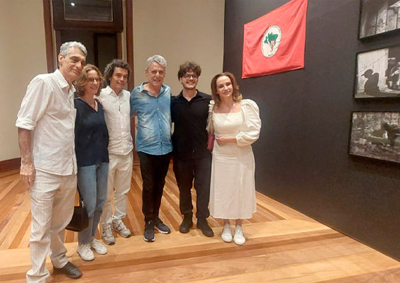 A imagem mostra um grupo de seis pessoas (entre elas o cantor Chico Buarque, a diretora-geral do CCJF, o curador e os artistas das exposições) posando na exposição Lona Preta: o MST no Olhar de Francisco Proner. O espaço tem paredes escuras e piso de madeira clara. Eles estão sorrindo e usam roupas casuais, predominantemente em tons claros. Ao fundo, há uma parede com fotografias em preto e branco e uma bandeira vermelha com um símbolo verde e branco. A expressão das pessoas sugere satisfação e camaradagem.