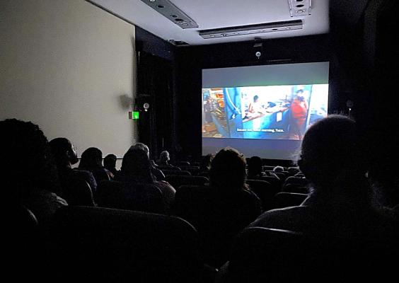 A imagem mostra uma sala de cinema escura com várias pessoas sentadas assistindo a um filme. A tela à frente exibe uma cena colorida de um ambiente interno, onde há duas pessoas e uma legenda em inglês na parte inferior, que diz: “Excuse me, good morning, Tuca.” As cabeças das pessoas na plateia aparecem em silhueta contra a luz da tela. O ambiente transmite uma atmosfera tranquila e imersiva, típica de uma sessão de cinema.