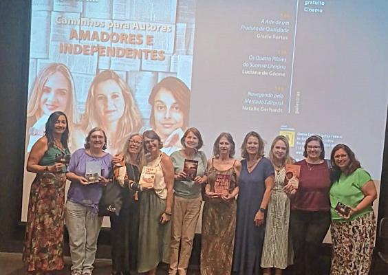Onze mulheres posando para uma foto em frente a uma tela de projeção em um auditório. Na tela, há um cartaz com o título "Caminhos para Autores Amadores e Independentes" e fotos de três palestrantes. O evento parece ser uma palestra ou oficina para escritores, com informações sobre os temas e nomes das palestrantes: Gisele Fortes, Luciana de Gnone e Natalie Gerhardt. As participantes na foto seguram livros, provavelmente obras próprias, indicando um encontro de autores independentes ou lançamento literário.