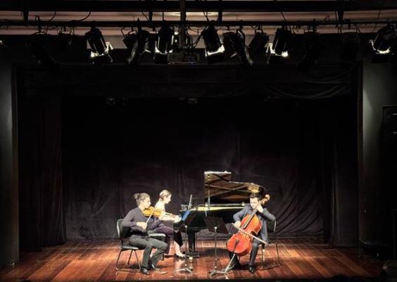 A imagem mostra três músicos em um palco de teatro com iluminação focada sobre eles, cercados por cortinas pretas. À esquerda, um violinista toca seu instrumento; ao centro, uma pianista sentada ao piano; e à direita, um violoncelista com seu violoncelo. Todos estão sentados em cadeiras e vestidos em roupas formais escuras. O chão do palco é de madeira, refletindo a luz suavemente. As luzes no teto estão direcionadas para o trio, criando um ambiente intimista para a performance musical.