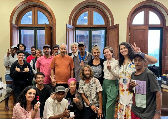 A imagem mostra um grupo de pessoas reunidas em uma sala com grandes janelas arqueadas. Elas sorriem e fazem gestos de paz ou descontração, transmitindo alegria e união. A diversidade do grupo é evidente, com pessoas de diferentes idades e estilos. Algumas seguram objetos simples, como uma vela em formato de número 2. O ambiente parece acolhedor, com piso de madeira e uma atmosfera de celebração ou encontro especial.