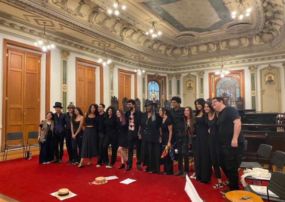 A imagem mostra um grupo de jovens, todos vestidos com roupas pretas, em sala com enorme tapete vermelho, portas em madeira, parede e teto com adornos de época. Eles estão em pé, sorrindo.