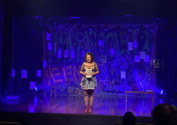 A imagem mostra uma mulher em pé no centro de um palco durante uma apresentação teatral. Ela veste um vestido curto azul estampado e sandálias vermelhas, segurando um objeto em suas mãos. O cenário ao fundo possui grafites coloridos, palavras escritas e papéis pendurados, criando um ambiente urbano e artístico. O palco é iluminado por luzes azuladas, contribuindo para uma atmosfera dramática e envolvente. Há uma bancada e um banco simples como parte dos elementos cênicos.
