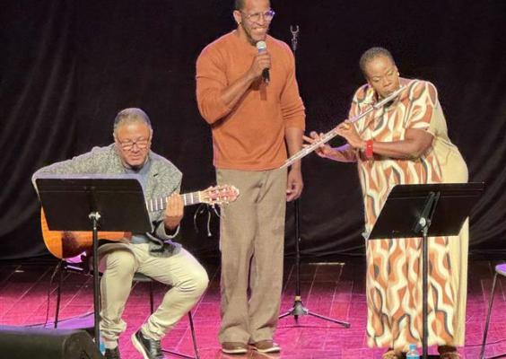 A imagem mostra três músicos se apresentando em um palco. À esquerda, um homem toca violão sentado, vestindo óculos, blusa cinza e calça clara, com uma partitura à sua frente. No centro, outro homem em pé segura um microfone, vestindo uma blusa marrom e calça bege, aparentando estar cantando. À direita, uma mulher toca flauta transversal, usando um vestido longo estampado e acessórios vermelhos. O fundo é preto e o palco tem um tom avermelhado, destacando os artistas e criando uma atmosfera intimista.