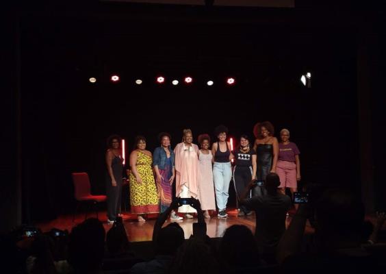 A imagem mostra um grupo de pessoas no palco de um teatro. O palco está iluminado por luzes vermelhas e brancas no fundo, enquanto o grupo posa para uma foto. São oito pessoas ao centro, vestindo roupas diversas e estilosas, com um público na plateia capturando o momento com câmeras ou celulares. A cena transmite uma atmosfera de celebração ou conclusão de um evento, como uma apresentação artística, uma performance ou uma cerimônia.