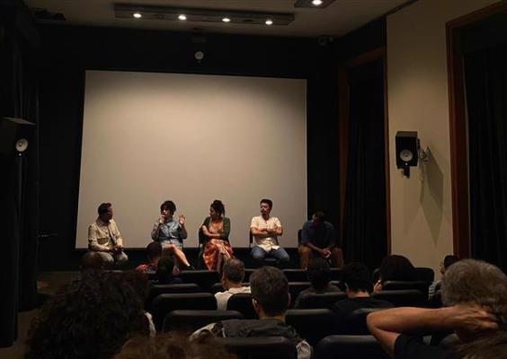 A imagem retrata uma sessão de conversa em uma sala de cinema ou auditório. Cinco pessoas estão sentadas à frente, participando de um painel ou discussão. O público, visível em primeiro plano, assiste atentamente. O cenário é iluminado suavemente, com um grande telão branco ao fundo, sugerindo uma exibição seguida de debate, como acontece em eventos culturais ou mostras de cinema.
