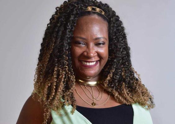 Esta imagem mostra uma mulher negra sorrindo, vestindo uma blusa clara com detalhes em preto e adornada com colares dourados. Seu cabelo é longo, volumoso e cacheado, com mechas claras nas pontas. Ela também usa uma tiara dourada que complementa seu visual. O fundo neutro e a iluminação suave destacam seu rosto expressivo e seu sorriso caloroso.