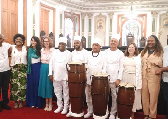 A imagem mostra um grupo de pessoas posando juntas em um ambiente elegante, com arquitetura clássica, incluindo colunas e detalhes decorativos nas paredes. Algumas pessoas vestem roupas brancas tradicionais e estão ao lado de tambores, sugerindo uma conexão com práticas culturais ou religiosas. Outras estão em trajes formais ou coloridos, sorrindo. Ao fundo, há janelas com vitrais e uma atmosfera de celebração ou cerimônia.