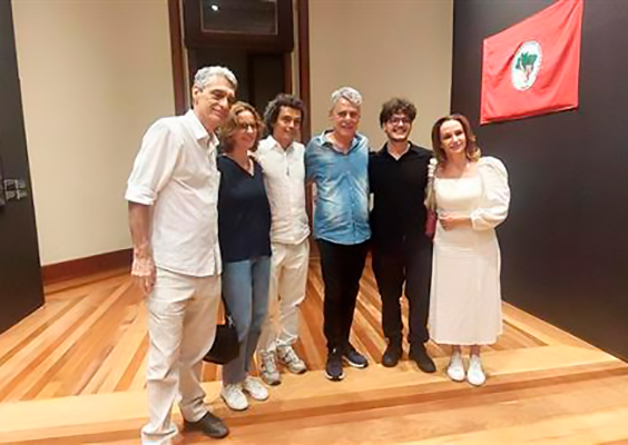 A imagem mostra seis pessoas em uma sala de exposições do Centro Cultural Justiça Federal (CCJF), com piso de madeira e a bandeira vermelha do MST ao fundo. O evento é a inauguração das exposições "Infinitos - Pinturas Recentes de Maria Lynch", "Espaços Quânticos", de César Oiticica Filho, e "Lona Preta: o MST no Olhar de Francisco Proner". Estão presentes Evandro Salles, Simone Schreiber, César Oiticica Filho, Chico Buarque, Francisco Proner e Carol Proner, em clima de celebração cultural.