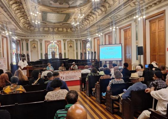 A imagem mostra um evento em uma sala histórica com arquitetura rica, teto decorado e vitrais ao fundo. Pessoas estão sentadas em bancos de madeira, assistindo a uma mesa de debate composta por quatro participantes, com um banner vermelho na frente indicando "Intolerância Religiosa". Um palestrante em traje branco está em um púlpito à esquerda. À direita, há um projetor exibindo o tema do evento. O ambiente tem detalhes clássicos e iluminação com lustres.