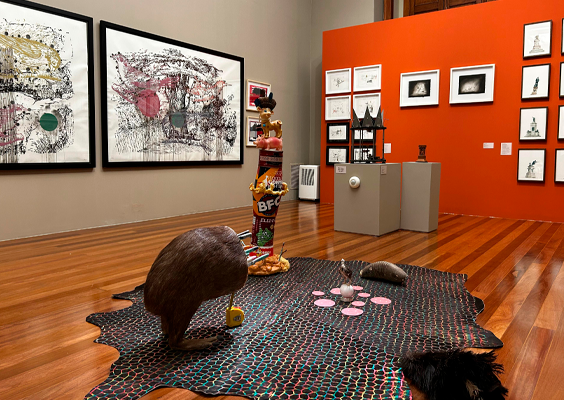 Interior de galeria de arte. No centro, há uma instalação artística composta por tapete com padrão geométrico, esculturas de animais, como um pássaro marrom, um tatu e a metade inferior de uma capivara com diversas facas cravadas no local de onde seria a cabeça, além de outros objetos inusitados. Ao fundo, quadros grandes com arte abstrata, pendurados na parede cinza. À direita, uma parede laranja exibe quadros menores em arranjo ordenado. O piso de madeira polida dá ao espaço um ar sofisticado e acolhedor.