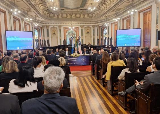 A imagem mostra um congresso em um salão nobre com arquitetura clássica, teto ornamentado e vitrais ao fundo. O evento reúne um grande público, composto por profissionais de terno e trajes formais, assistindo a uma mesa de palestrantes. Duas telas exibem o título "I Congresso - Nova Arquitetura da Recuperação da Saúde - Impactos do Tema 1234". Bandeiras oficiais estão posicionadas ao centro, e uma pessoa discursa em um púlpito à esquerda.
