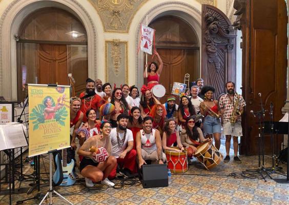 Grupo animado de pessoas vestidas com roupas vermelhas, brancas e acessórios carnavalescos posa sorridente em um saguão com arquitetura clássica. No centro, um cartaz anuncia 'Bloco dos Inconfidentes'. Instrumentistas seguram saxofone e tambor, enquanto uma mulher no alto levanta um estandarte do bloco.
