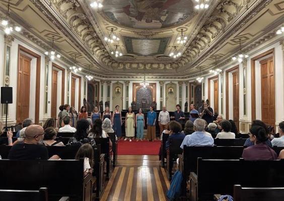 Coral de cantores se apresenta no centro da Sala de Sessões do CCJF, enquanto a plateia assiste sentada em bancos de madeira. A arquitetura sofisticada e os trajes leves dos artistas criam um contraste harmonioso entre tradição e modernidade.