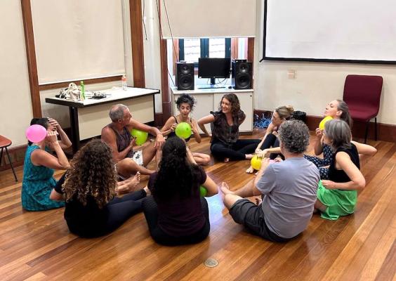Grupo de pessoas sentadas em círculo em uma sala com piso de madeira, segurando balões coloridos. O ambiente tem uma tela de projeção, caixas de som e uma mesa com objetos ao fundo. Todos parecem engajados em uma atividade interativa, com expressões de concentração e diversão.