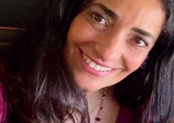 Na imagem, Claudia Domingues, responsável pelo Setor de Música do CCJF, uma mulher de cabelos longos e cacheados sorri. Ela usa um colar com pedras e uma blusa roxa. Seu olhar transmite simpatia e alegria. O fundo está desfocado, destacando seu rosto iluminado.