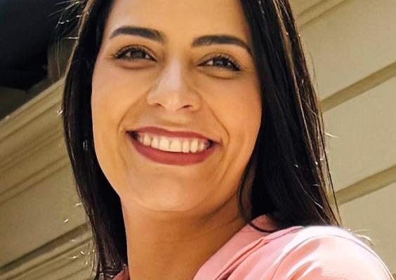 Na imagem, Klara Freire, bibliotecária do CCJF, uma mulher de cabelos castanhos longos, sorri para a câmera. Ela veste um blazer rosa sobre uma blusa branca. O fundo mostra uma parede clara com detalhes arquitetônicos e luz natural entrando pelo topo da imagem.