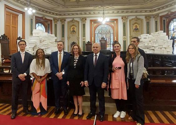 A imagem mostra um grupo de oito pessoas posando para uma foto em um salão com arquitetura clássica, repleto de detalhes ornamentais. O fundo possui um grande número de caixas brancas empilhadas e vitrais decorativos. As pessoas estão vestidas com trajes formais e semiformais. Entre elas, há homens de terno e gravata, e mulheres com vestidos e roupas casuais elegantes. O ambiente tem um ar solene, sugerindo uma visita institucional ou evento oficial.