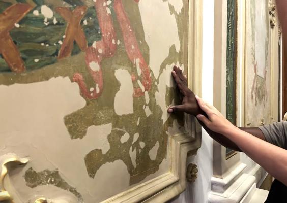 Duas pessoas tocam uma pintura antiga e desgastada em uma parede ornamentada, provavelmente em um museu ou prédio histórico. Uma das mãos é de pele escura e está em contato direto com a pintura, enquanto a outra mão, de pele clara, repousa suavemente sobre a mão anterior, sugerindo orientação ou apoio tátil. A parede ao fundo é decorada com molduras e outras obras de arte.