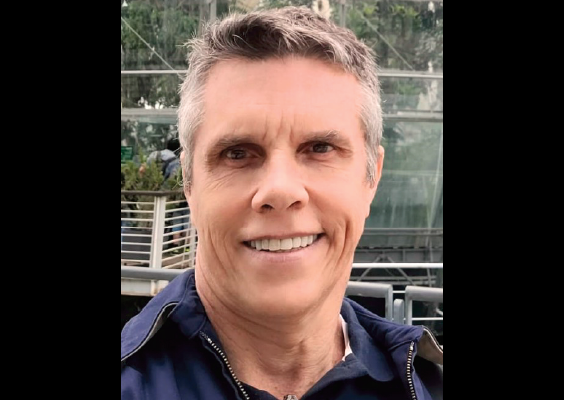 Homem de cabelos curtos e grisalhos sorrindo para a câmera em ambiente externo. Ele veste uma jaqueta azul sobre uma camisa polo e está em frente a uma estrutura de vidro com vegetação interna, que lembra uma estufa ou construção moderna com plantas. Ao fundo, há uma passarela metálica e outras pessoas ao redor.