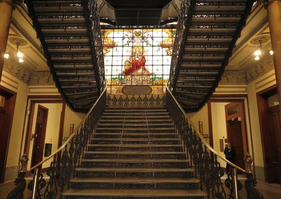 A imagem mostra uma escadaria grandiosa e simétrica em um prédio histórico. A estrutura tem corrimãos e grades de ferro ornamentado, e os degraus levam a um vitral colorido no topo, representando uma figura feminina. O ambiente é ricamente decorado, com colunas douradas, detalhes em gesso no teto e portas de madeira. Há uma pessoa uniformizada no canto direito, possivelmente um segurança.
