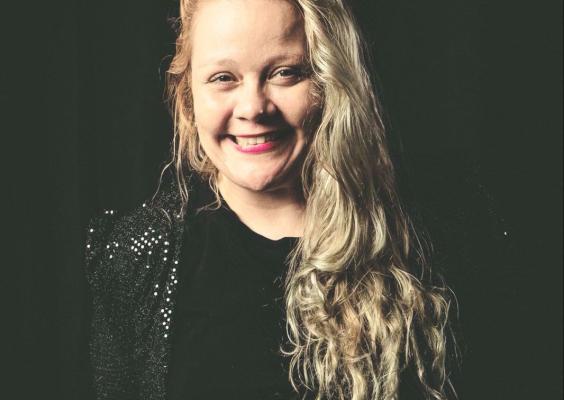 Mulher loira de cabelos longos e ondulados, sorrindo para a câmera em um fundo escuro. Ela veste uma blusa preta com ombreiras brilhantes de lantejoulas. A iluminação destaca seu rosto e cabelo, criando um contraste com o fundo preto, dando um ar de retrato artístico.