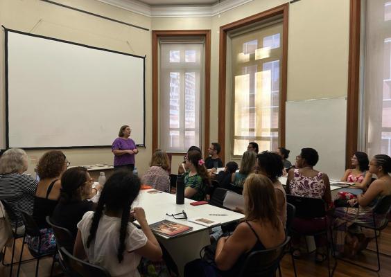Sala de aula com janelas grandes e iluminação natural. Uma mulher em pé, vestindo blusa roxa, fala para um grupo atento de pessoas sentadas ao redor de mesas. Os participantes têm cadernos, livros e garrafas sobre as mesas. Ao fundo, há um quadro branco e uma grande tela de projeção.