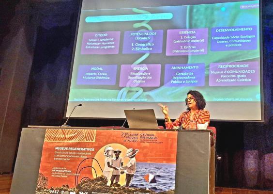 Imagem de uma palestra sobre “Museus Regenerativos” em um auditório. Uma mulher com cabelo cacheado, usando blusa vermelha estampada, está sentada à mesa com um microfone, notebook e xícara. Atrás dela, um slide apresenta conceitos como “O Todo”, “Essência” e “Reciprocidade”. Na frente da mesa, há um cartaz do evento “23ª Semana Nacional de Museus” com a frase “Construindo futuros sustentáveis com comunidades em rápida transformação”.