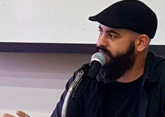 Homem de barba cheia e camisa escura fala ao microfone durante uma mesa ou palestra. Ele usa boina preta e camiseta com estampa da lua, gesticulando com a mão esquerda enquanto segura o microfone com a direita. Ao fundo, parede branca e parte de uma tela de projeção são visíveis. A expressão facial é séria e concentrada, indicando envolvimento com o tema abordado.