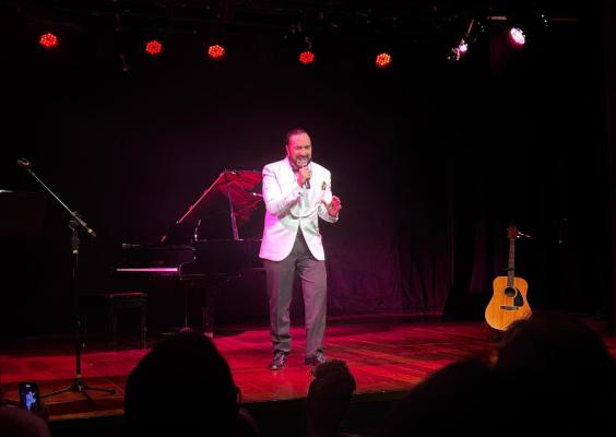 Helcio Hime se apresenta no palco usando paletó branco e camisa escura, segurando um microfone e cantando com expressão intensa. Ao fundo, há um piano de cauda e um violão acústico apoiado em pé. A iluminação é predominantemente vermelha e púrpura, criando clima intimista. Público assiste à performance; uma pessoa fotografa com o celular no canto inferior esquerdo.