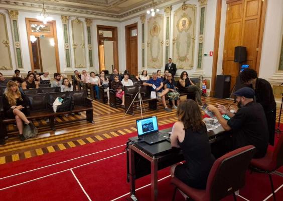 Na imagem, a Sala de Sessões recebe o evento com plateia atenta. Três pessoas estão na mesa de frente, com notebooks e materiais impressos; uma delas fala enquanto outra mostra um objeto. Ao fundo, público variado observa a apresentação. Cadeiras de madeira, tapete vermelho e iluminação pendente compõem o ambiente sofisticado e histórico.
