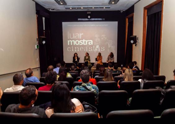 Sessão de debate na Sala de Cinema com projeção da tela ao fundo exibindo os dizeres "Centro Cultural Justiça Federal e Coletivo Luar apresentam: luar mostra cineastas". Quatro pessoas estão sentadas à frente do público, participando de uma mesa de conversa. A plateia, composta por diversas pessoas, assiste atentamente.