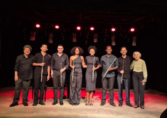 Grupo de músicos posando no palco após apresentação. Sete pessoas estão na foto, todas vestindo roupas pretas (exceto uma pessoa à direita, que veste uma blusa verde claro). Cada um segura uma flauta. O cenário está iluminado com luzes vermelhas ao fundo e há uma atmosfera de concerto. As pessoas são de diferentes etnias e idades, e todas sorriem para a câmera. À esquerda, o grupo está composto por um homem mais velho, dois homens jovens, duas mulheres, e três homens de meia-idade.