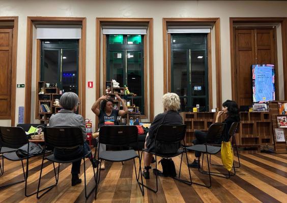 Três pessoas estão sentadas em cadeiras dispostas em semicírculo dentro de uma sala com piso de madeira e grandes janelas. À frente delas, uma mulher no centro fala gesticulando com as mãos, enquanto as outras escutam atentamente. Atrás dela, há estantes com livros e brinquedos. 