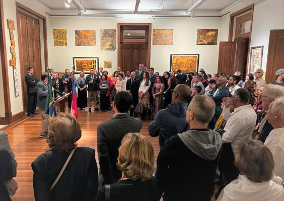 A imagem mostra uma sala de exposições cheia de pessoas, durante uma cerimônia de abertura. À esquerda, um homem fala ao microfone ao lado das bandeiras do Brasil e do Chile. O público, em pé, observa com atenção. Nas paredes, há quadros com paisagens urbanas e cores quentes. O ambiente tem piso de madeira e iluminação direcionada, transmitindo um clima solene e artístico.