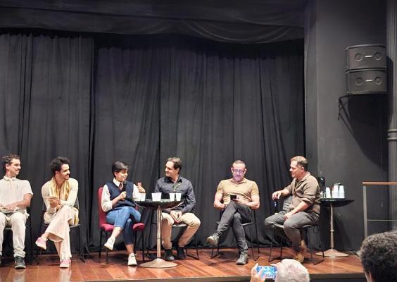 A imagem mostra um painel com seis pessoas sentadas em cadeiras no palco de um teatro, com cortinas pretas ao fundo. Três homens e uma mulher ao centro conversam animadamente, um deles segurando um microfone. Nas extremidades, dois homens observam. Há copos sobre mesas pequenas e uma pessoa do público tira uma foto na frente do palco.