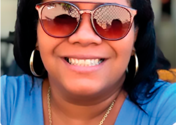 Mulher com cabelos curtos e escuros, usando óculos de armação grossa azul e dourada. Ela sorri levemente para a câmera e veste uma camisa azul clara com detalhes vermelhos na gola. Ao fundo, uma imagem desfocada.