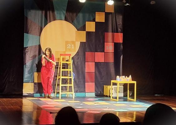 A atriz Bia de Queiroz , vestindo um macacão vermelho, apresenta um monólogo em um palco colorido, decorado com painéis geométricos numerados e um cenário lúdico. Ela interage com uma escada amarela e uma mesa com copos e garrafas, enquanto o público assiste em um teatro escuro.