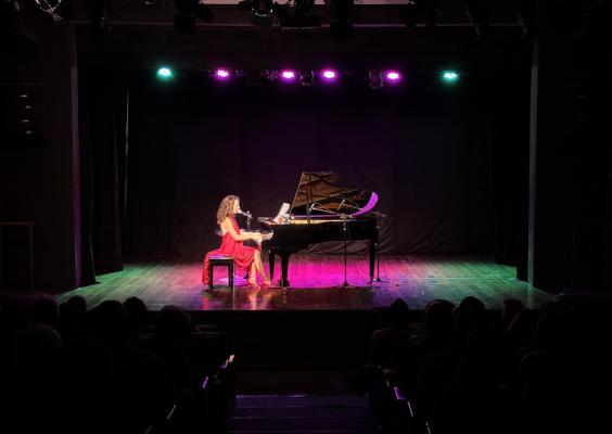 Julie Wein se apresenta em um palco iluminado por luzes roxas e verdes, usando um vestido vermelho. Ela toca piano enquanto canta ao microfone, com o público assistindo em um teatro escuro.