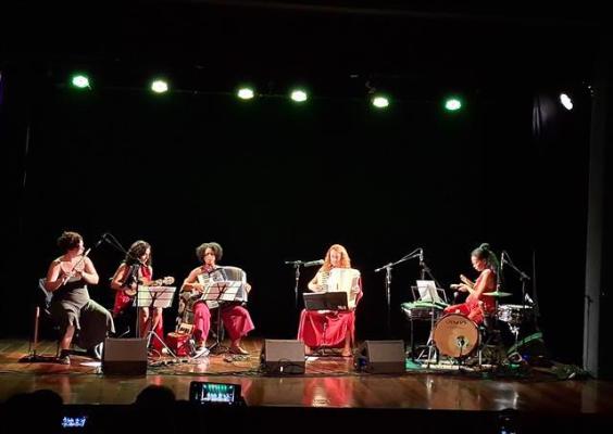 Cinco musicistas se apresentam em um palco iluminado por luzes verdes. Todas estão sentadas, tocando instrumentos como violino, cavaquinho, violão, sanfona e bateria. Elas usam roupas em tons de vermelho e estão concentradas na performance. O fundo do palco é escuro, destacando ainda mais as artistas sob os refletores. A plateia aparece em silhueta, com celulares gravando o show no teatro escuro.