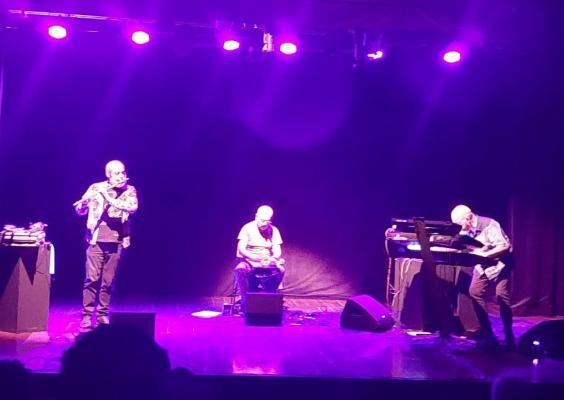 Apresentação dos musicistas Malta, Suzano e Meirelles em um palco escuro, iluminado por luzes roxas. Os três estão no palco: o primeiro, à esquerda, toca flauta e veste uma jaqueta estampada sobre camiseta preta e calça escura; o segundo, no centro, está sentado, tocando um instrumento de percussão, usando camiseta clara e calça escura; o terceiro, à direita, toca teclado, vestindo uma camisa azul estampada e calça escura. Enquanto o público assiste no teatro escuro.
